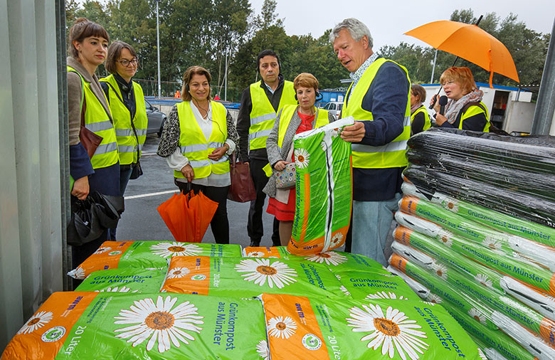 Huit personnes en gilet jaune de sécurité, la femme la plus à droite avec un parapluie orange, regardent un homme qui soulève un sac de compost vert.